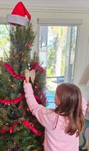 Picture of child with brown hair wearing a pink shirt looking at an ornament on a Christmas tree. The ornament is in the form of angel wings and has Andy's Angels written on it.